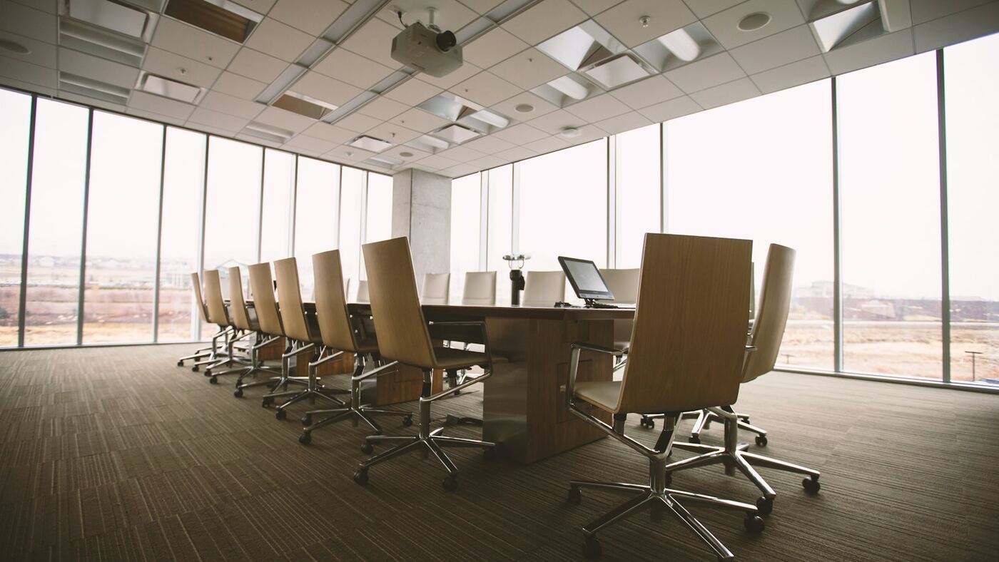 Empty modern conference room with a long wood table, rolling chairs, ceiling-mounted projector, and floor-to-ceiling windows