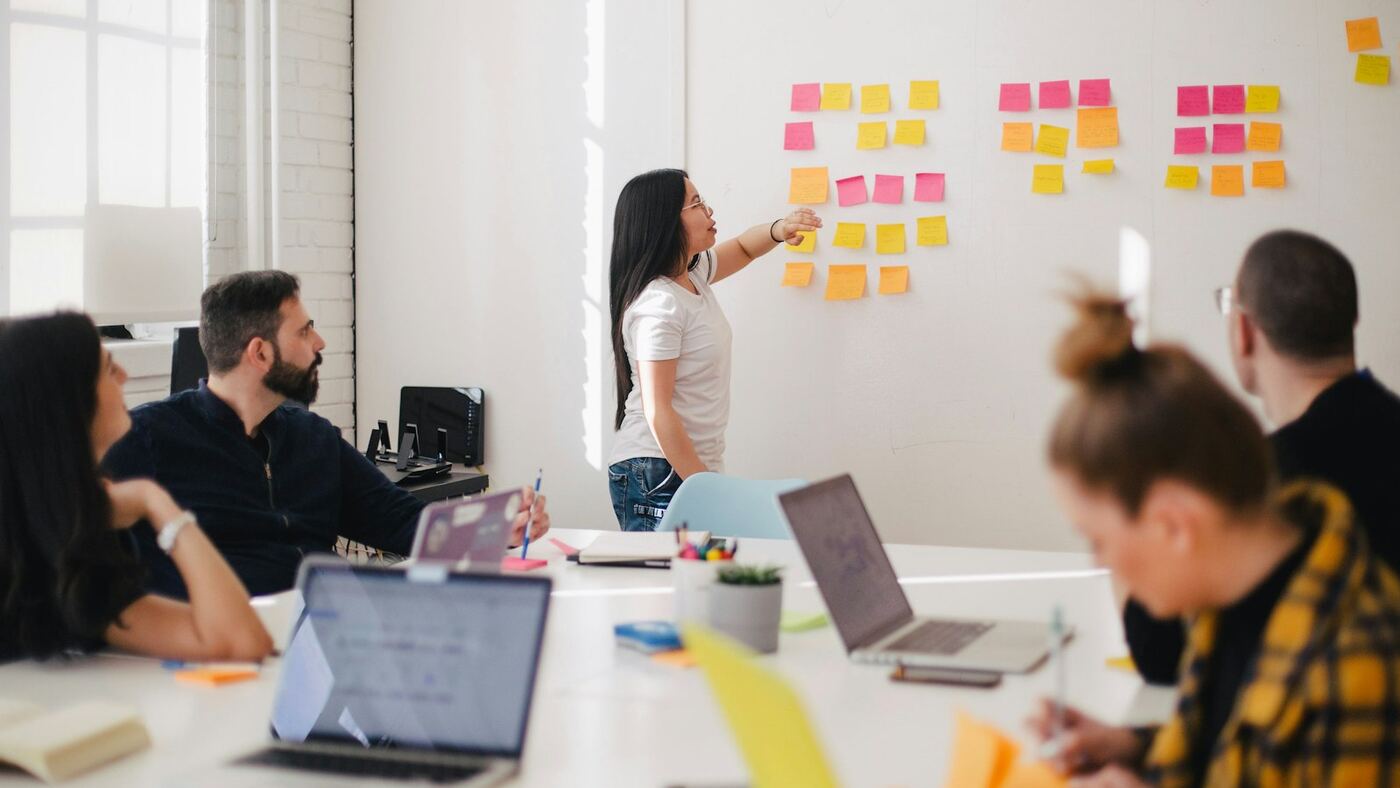 Team collaborating in a bright loft office around a table with laptops while a colleague presents sticky-note plans on the wall
