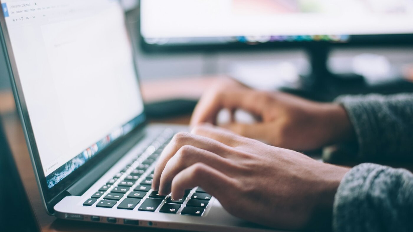 Hands typing on a silver laptop keyboard at a wooden desk with a second monitor softly blurred in the background